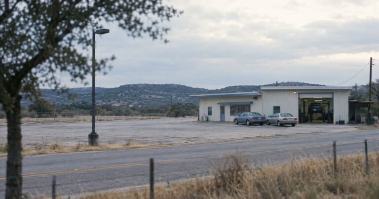 A small flat-roofed commercial building with one open roller door at the edge of a Texas town, two cars outside and an empty paved lot beside it, the hero image of our guide to checking if a car dealership is legit